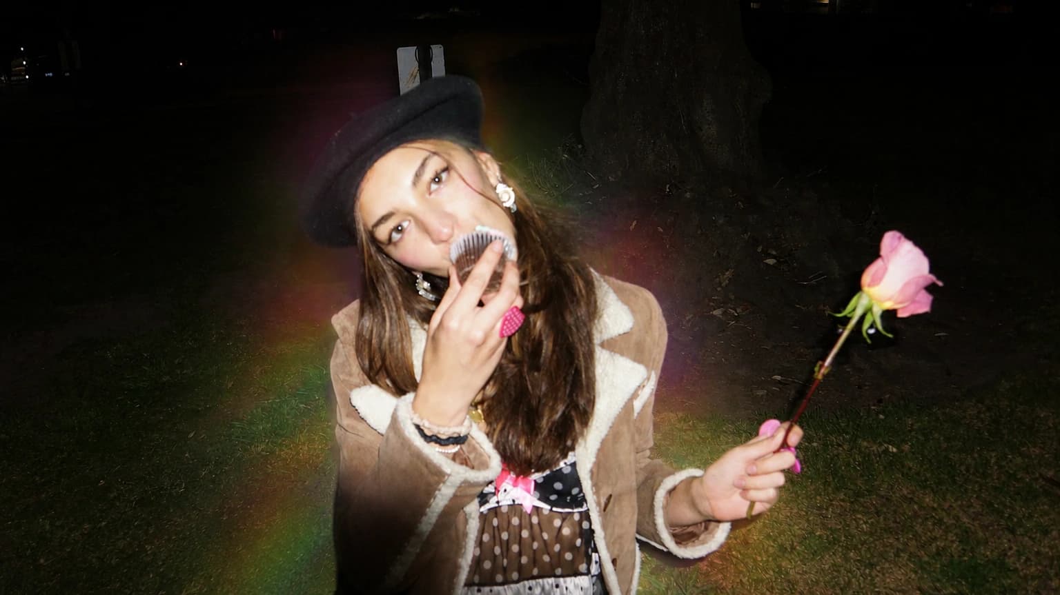 A woman in a black beret and shearling coat eating a cupcake at night, holding a pink rose