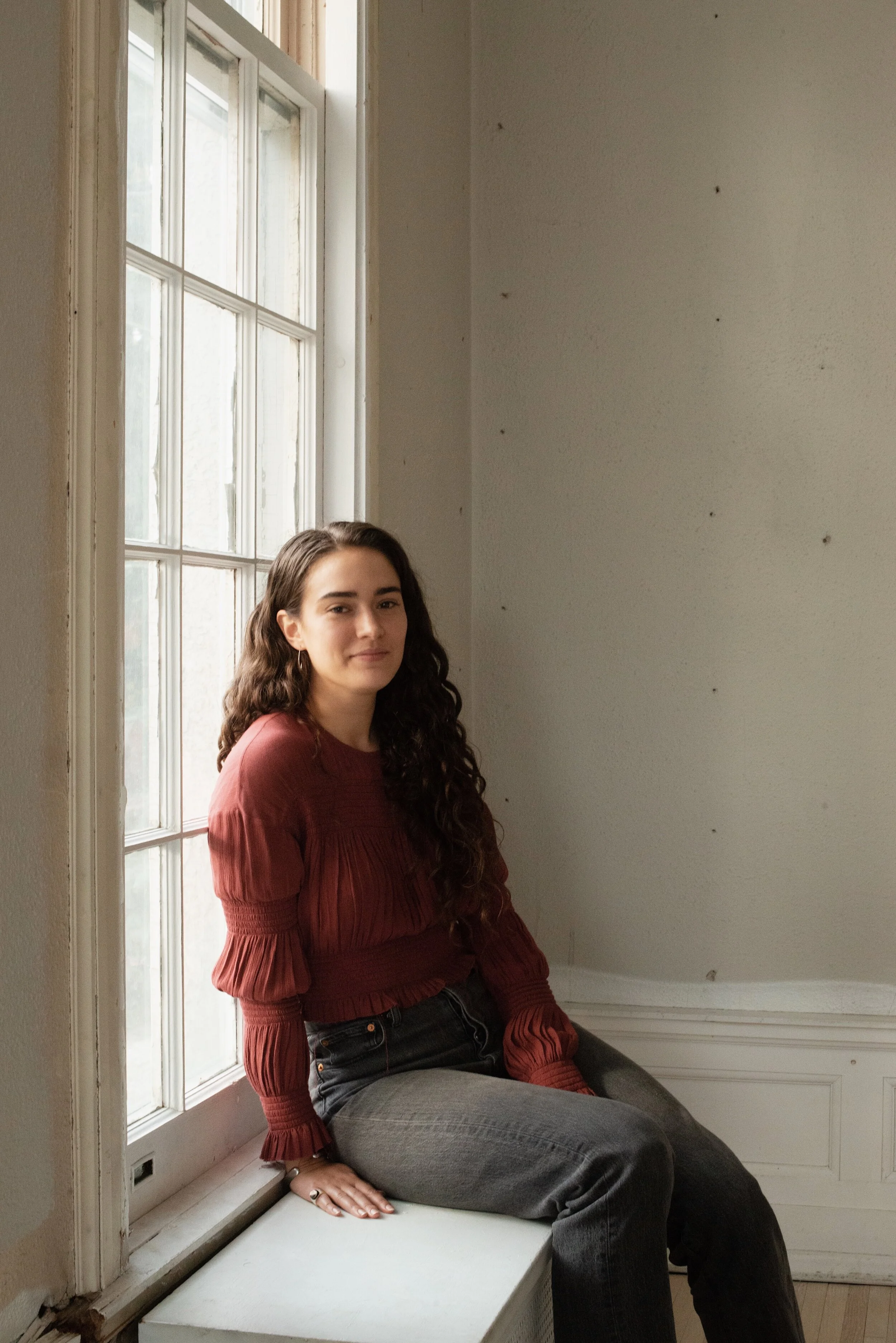 Lily Harris sitting on a windowsill in a rust-toned blouse, smiling softly